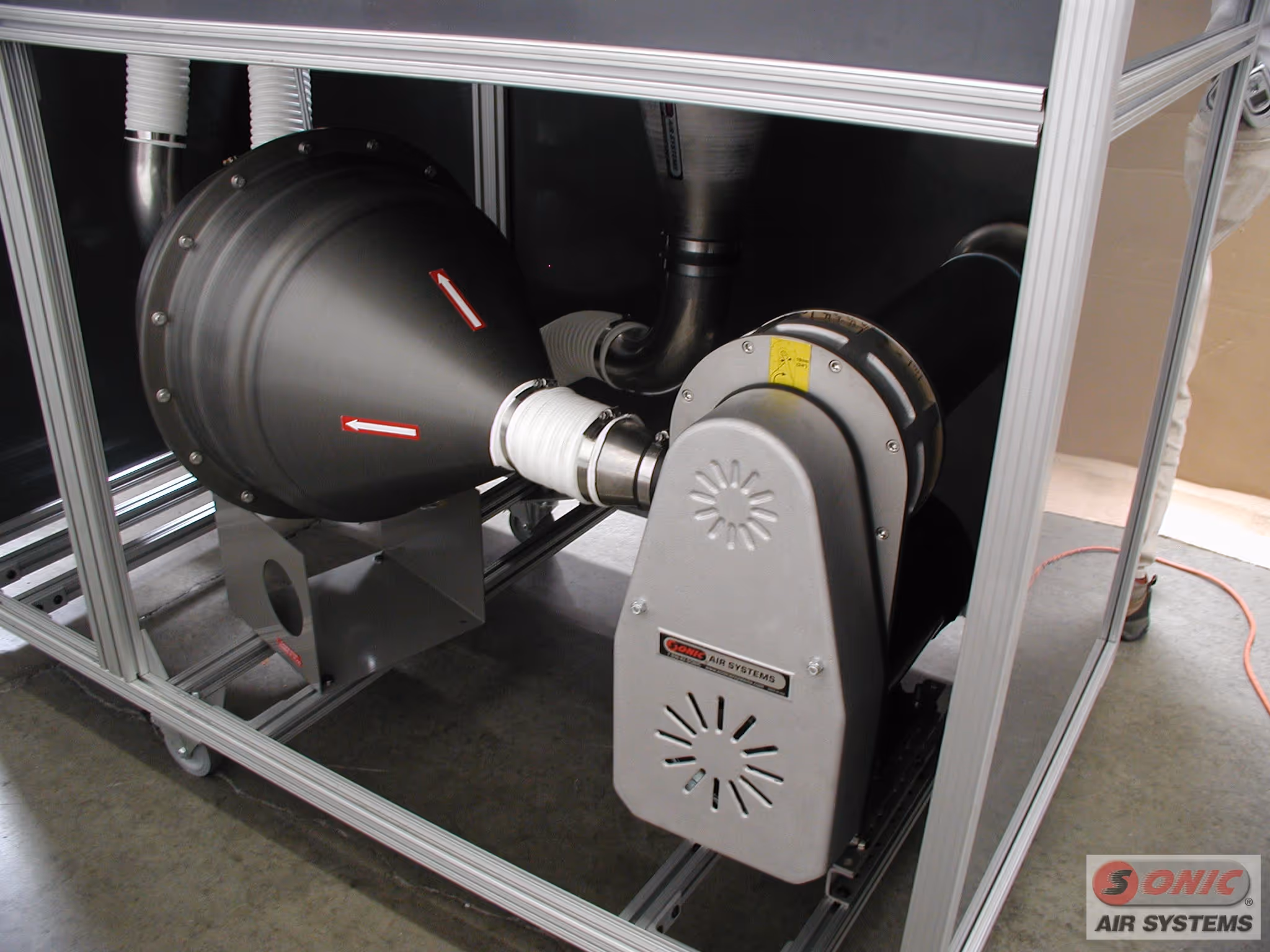 Primer plano de un conjunto de soplador centrífugo industrial y filtro de Sonic Air Systems, montado en un bastidor de aluminio, con carcasa cónica negra, flechas rojas de dirección y ducto flexible blanco.