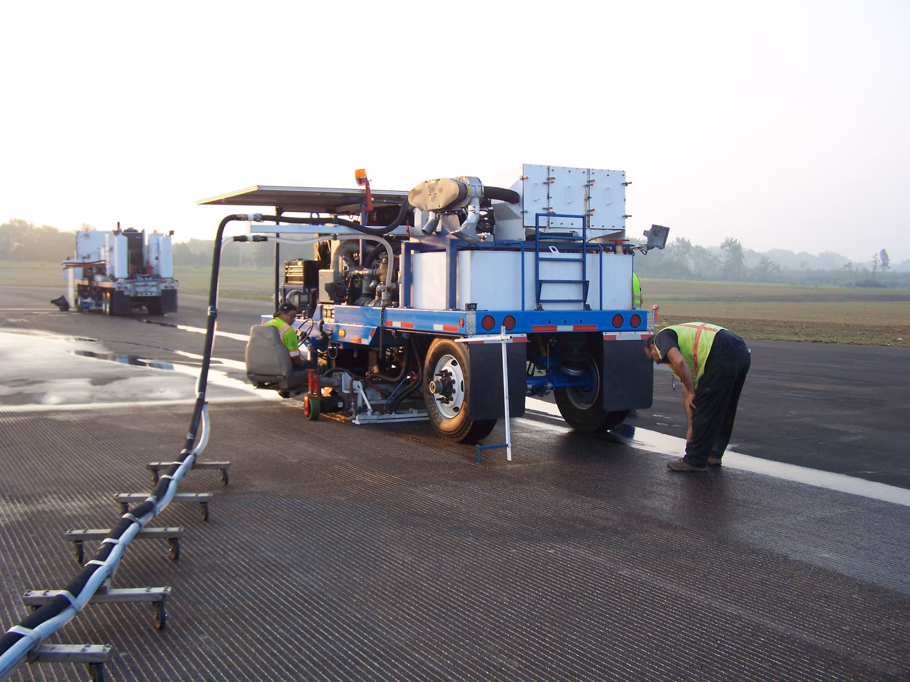 Un trabajador inspecciona la superficie mojada y ranurada de una pista de aeropuerto detrás de una máquina especializada de ranurado y chorro de agua conectada por líneas de suministro largas.