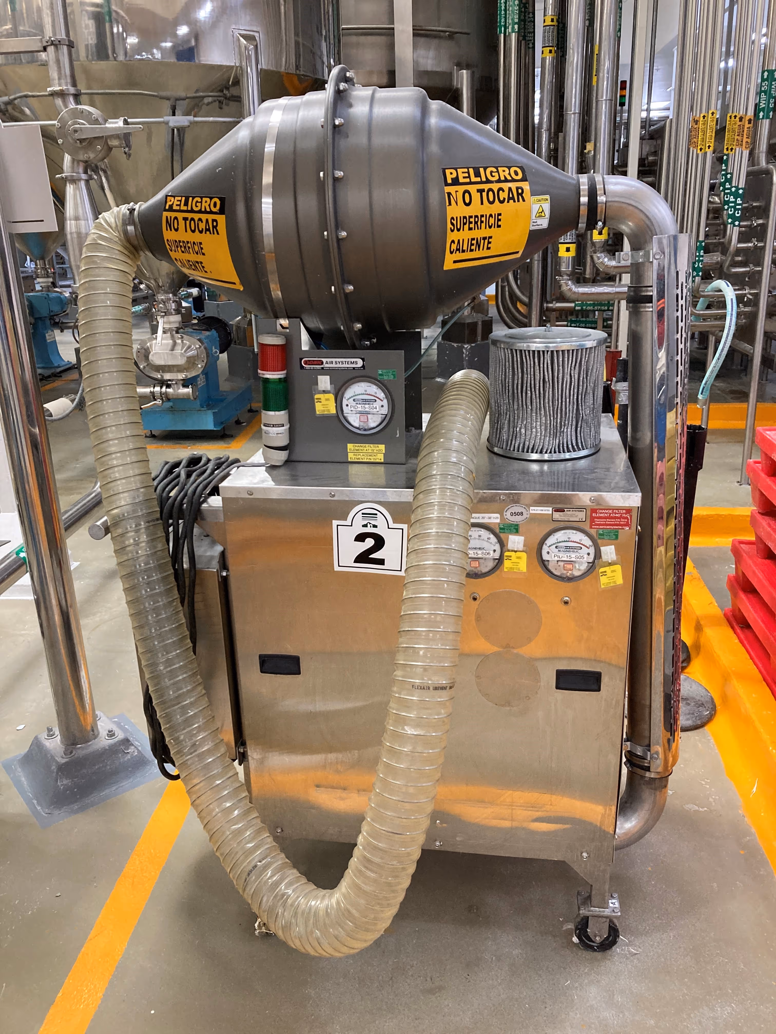 Unidad móvil de filtración de aire industrial con dos etiquetas de advertencia que dicen "PELIGRO NO TOCAR SUPERFICIE CALIENTE" y mangueras flexibles, sobre un piso de fábrica con franjas naranjas en una cervecería o planta de proceso.